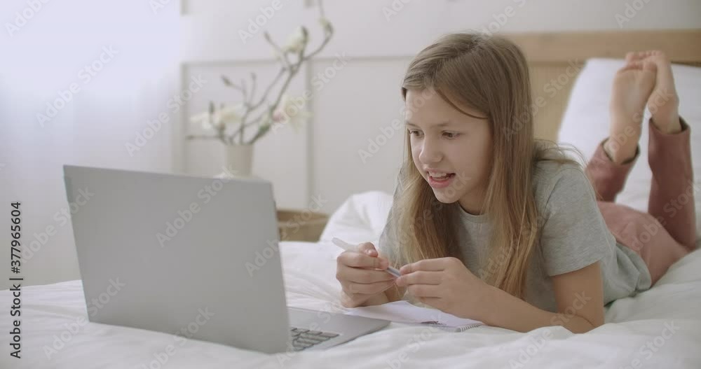 cheerful pupil girl is answering during online lesson by laptop, lying on bed and speaking, e-learning cheerful pupil girl is answering during online lesson by laptop, lying on bed and speaking, e-learning