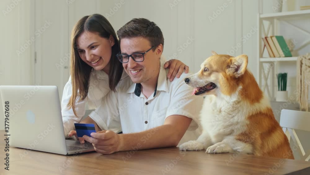 Cheerful Young Couple With a Dog Shopping Online While Sitting Home. Mutual Family's Purchases. Purchase Confirmation by the Internet. Binding a Card for Online Shopping Cheerful Young Couple With a Dog Shopping Online While Sitting Home. Mutual Family's Purchases. Purchase Confirmation by the Internet. Binding a Card for Online Shopping