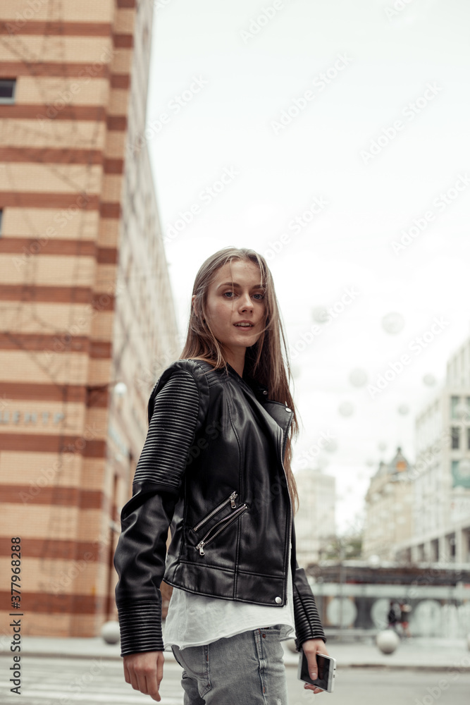 portrait girl with blonde hair on the city building background. Young woman dressed in black leather jacket, grey jeans and white shirt. Model walking and crossing a road