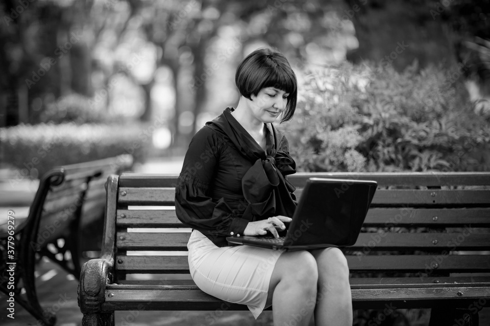 Fototapeta premium black and white portrait of a woman in business style, with a laptop, on a bench in the Park, working remotely in the field of Economics and accounting.