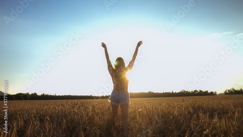 Happy woman walking on a wheat field with open arms at the bright sun.