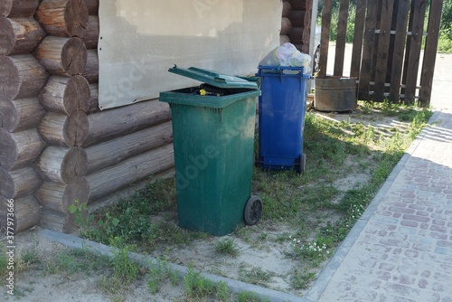 two full plastic trash cans stand on green grass and ground outside against a brown wooden wall © butus