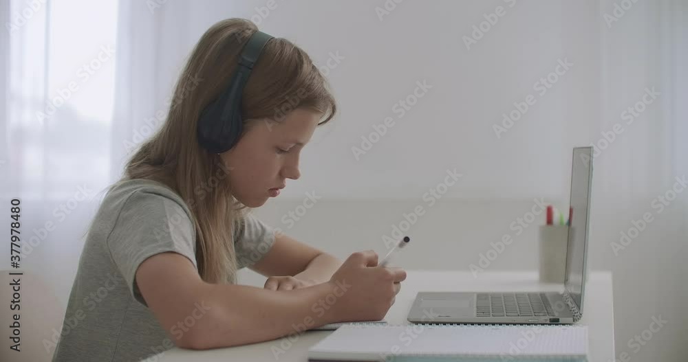 schoolgirl is doing homework sitting at table in her room, using laptop and headphones, writing in exercise book schoolgirl is doing homework sitting at table in her room, using laptop and headphones, writing in exercise book