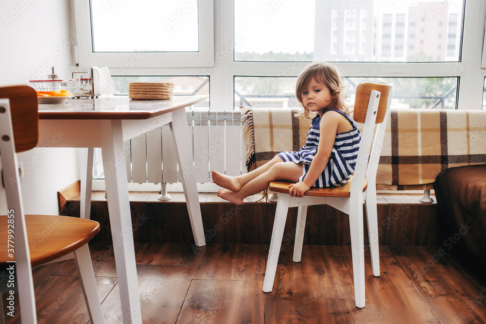 sad child sits alone on a chair in the kitchen. The girl looked down and sits on a stool at an empty table sad child sits alone on a chair in the kitchen. The girl looked down and sits on a stool at an empty table