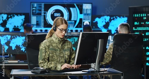 Caucasian female army officer in glasses and headset talking and working on computer in analytic secret base in military. Woman militarian official typing in dark office. Digital support. Surveillance photo