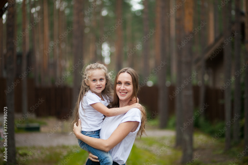 Fototapeta premium Happy smiling mother and her cute emotional little daughter child in white shirts and denim jeans are hugging and having fun outdoor in nature at countryside. Spending summer time with family.