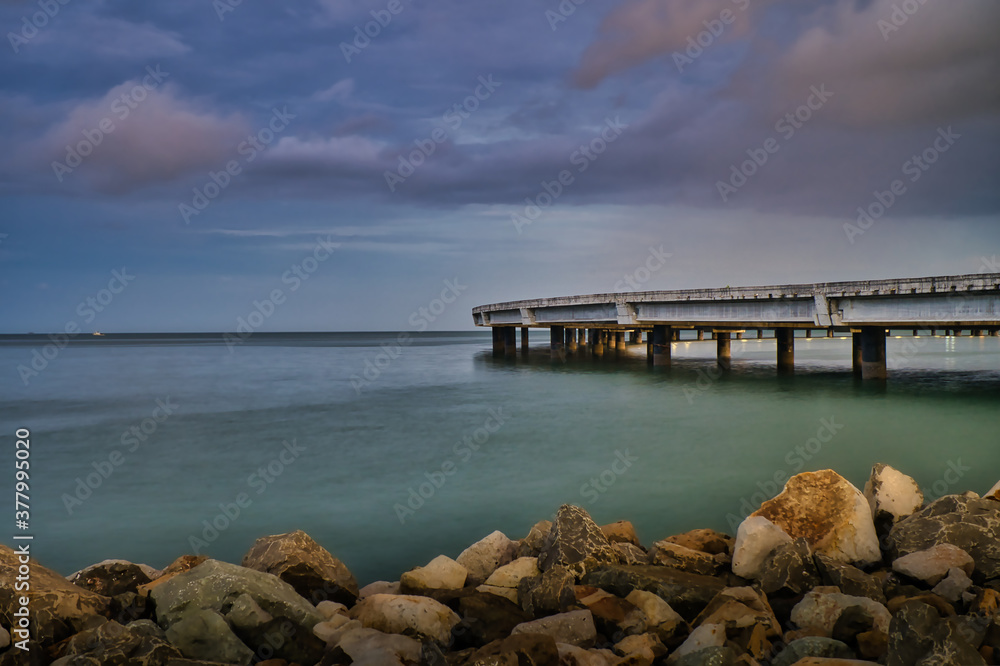 Eine menschenleere Brücke über das Meer in Panama, Steine im Vordergrund und lila Wolken, kurz vor einem Gewitter
