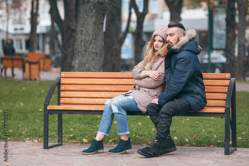 A guy and a girl are resting on a bench in an autumn park. A loving couple in jackets sits on a bench in the main park © Дмитрий Скорина
