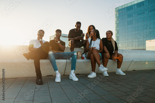 Group of young modern african black friends happily sitting together on the bench of the stairs in bright sunlight enjoying summer vacation