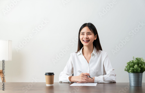 Smiley young Asian woman sitting at a wooden table with holding pen and document information on the desk. Look sideways and build imagination through creative ideas.