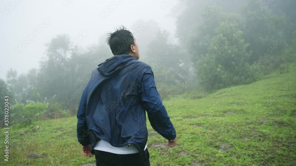 Slow motion of Asian man tourist in blue jacket walking in the rain against strong wind on the mountain. Male hiker hiking in raining day. Outdoor lifestyle, bad weather and travel vacation concept.