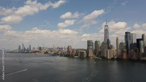 A bird's eye view over Upper Bay with water vessels moving by in the Hudson River below. The drone trucks right and pan left. It is a sunny day in New York and the West side of Manhattan is in view.