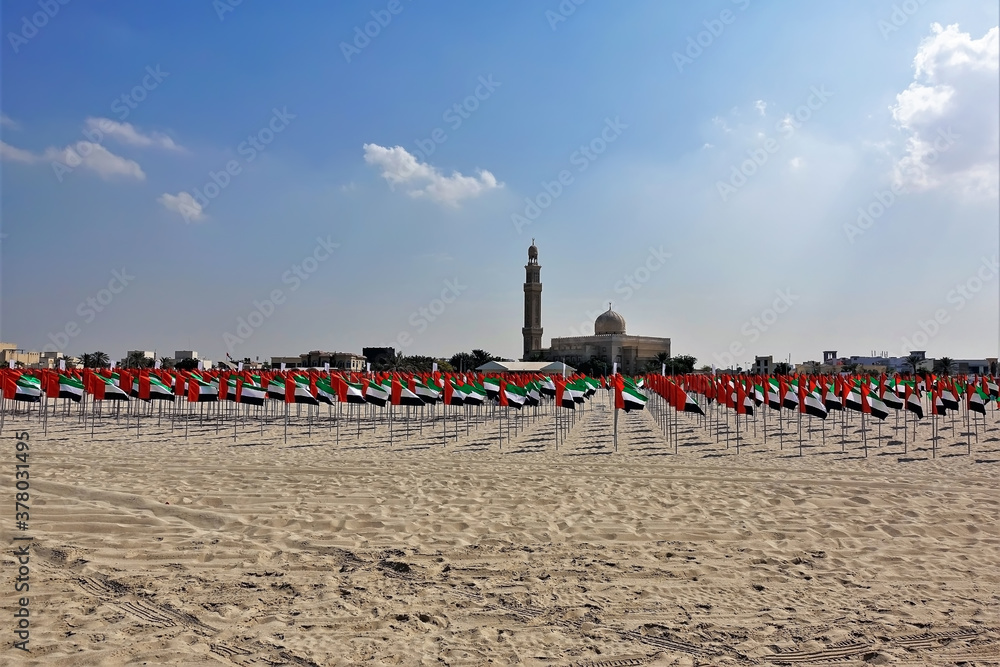 Celebration of the day of the state flag of the UAE. There are many rows of flags of black-red-white-green colors flying on the sand of the beach. Mosque against the blue sky. Dubai.