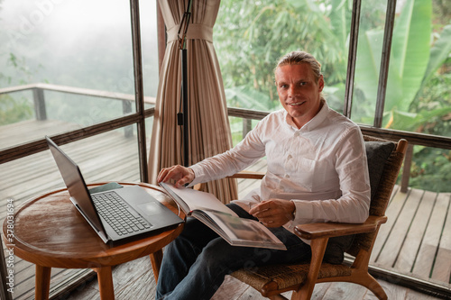 Happy smiling journalist stylish guy read a book  in a workplace in loft styled coworking, well dressed, sitting near window with view of garden lok at the camera © Yevhenii