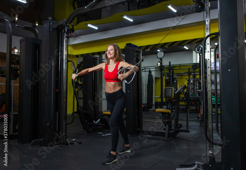 Smilling Fit woman flexing muscles on gym machine. Girl execute exercise with exercise-machine Cable Crossover in modern gym. Fitness and bodybuilding