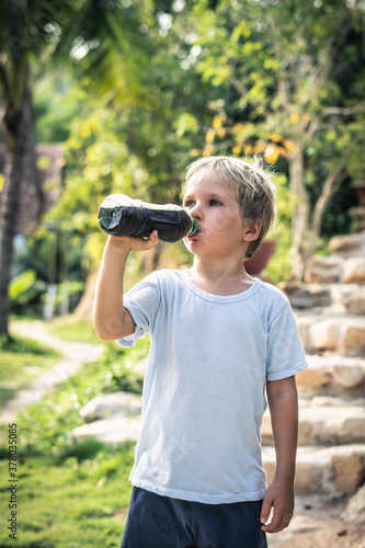 Front view of happy thirsty blond kid boy drinking dark tea from plastic bottle standing outside in summer park nature background. Healthy and unhealthy drinks concept. Stories vertical format