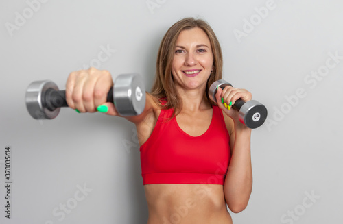 Slim attractive sports woman in a red sports top is exercising with dumbbells in her hands on a gray background. Healthy lifestyle, fitness concept