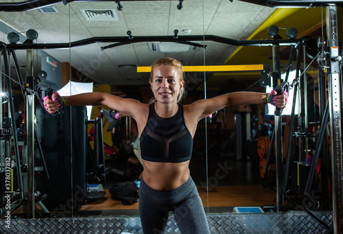 Slim Fit blonde woman flexing muscles on gym machine. Girl execute exercise with exercise-machine Cable Crossover in dark gym.