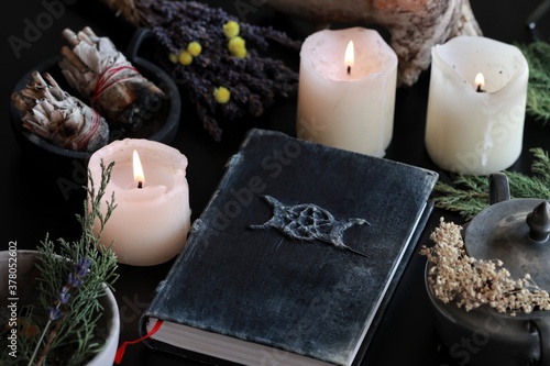 Wiccan witch altar with a hand made old looking book grimoire with triple moon symbol, and other various items - dried flowers, nature elements, burning candles, sage smudge sticks in black background photo