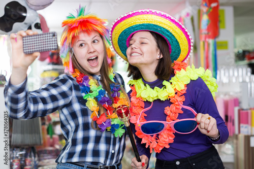 Comically dressed happy girls making funny selfies photo in festive accessories shop