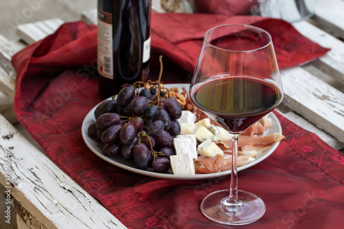 Wino czerwone i kieliszek .Talerz z prosciutto, serem brie i gorgonzolą, orzechami greckimi i winogronem.