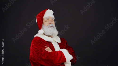 Waist up portrait of Santa Claus in traditional costume crossing arms and nodding head confidently in slow motion while looking at camera standing against black background