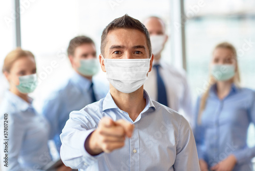 business, people and pandemic concept - businessman wearing face protective medical mask for protection from virus disease pointing finger to camera at office