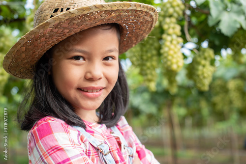 A cute girl in a hat and plaid shirt stood with her arms open in the garden, looking and smiling at the camera. The background is a vineyard. Asian children work hard to help the family business.