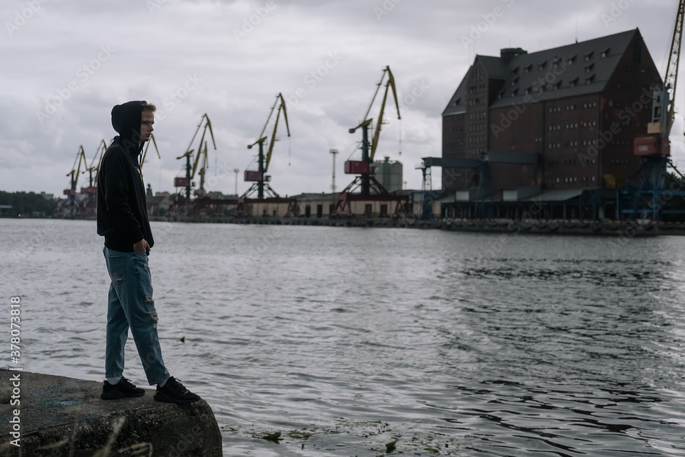A lonely unhappy anxious teenager stands by the river against the background of the port on a cloudy day