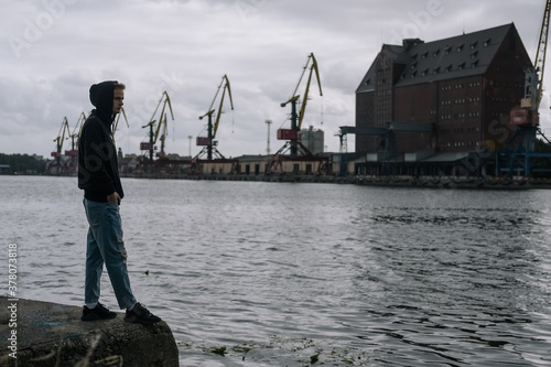 Obraz na płótnie A lonely unhappy anxious teenager stands by the river against the background of the port on a cloudy day w salonie