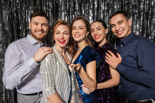 celebration, technology and holiday concept - happy friends taking picture by selfie stick at party