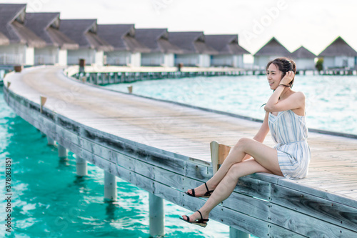 Beautiful young woman sitting on wooden pier by beautiful summer sun. Young Woman in white on a wooden beach pier. Girl enjoying vacation on wooden pier with water bungalow villa. Travel and Vacation.
