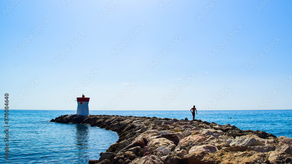 Silhouette of a young guy on the stones of the breakwater,  by the end of which stands a lighthouse.