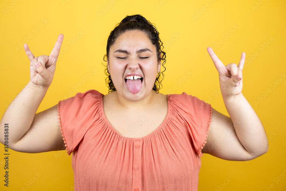 Young beautiful woman with curly hair over isolated yellow background shouting with crazy expression doing rock symbol with hands up