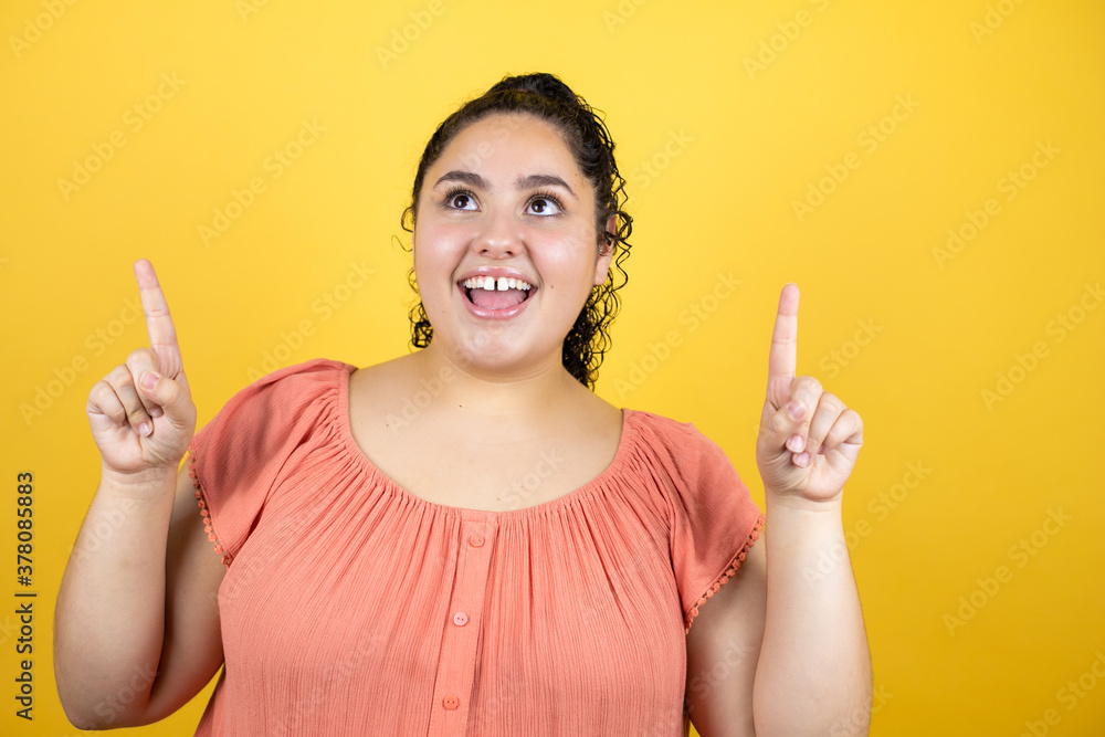 Young beautiful woman with curly hair over isolated yellow background amazed and surprised looking up and pointing up with fingers and raised arms Young beautiful woman with curly hair over isolated yellow background amazed and surprised looking up and pointing up with fingers and raised arms