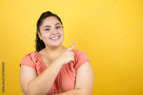 Young beautiful woman with curly hair over isolated yellow background smiling happy pointing with hand and finger to the side