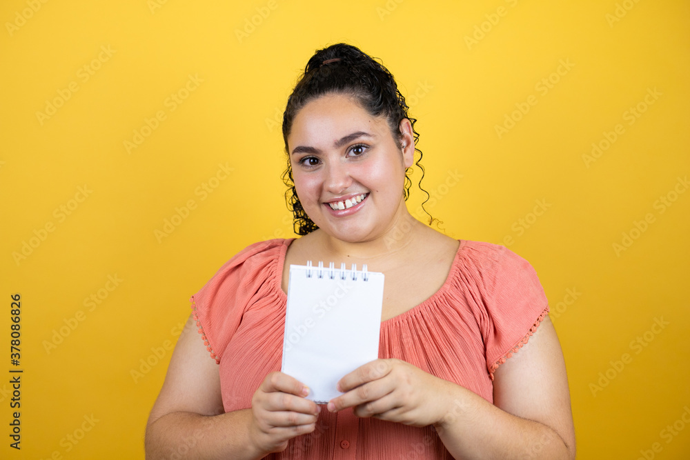 Young beautiful woman with curly hair over isolated yellow background smiling and showing blank notebook in her hand Young beautiful woman with curly hair over isolated yellow background smiling and showing blank notebook in her hand
