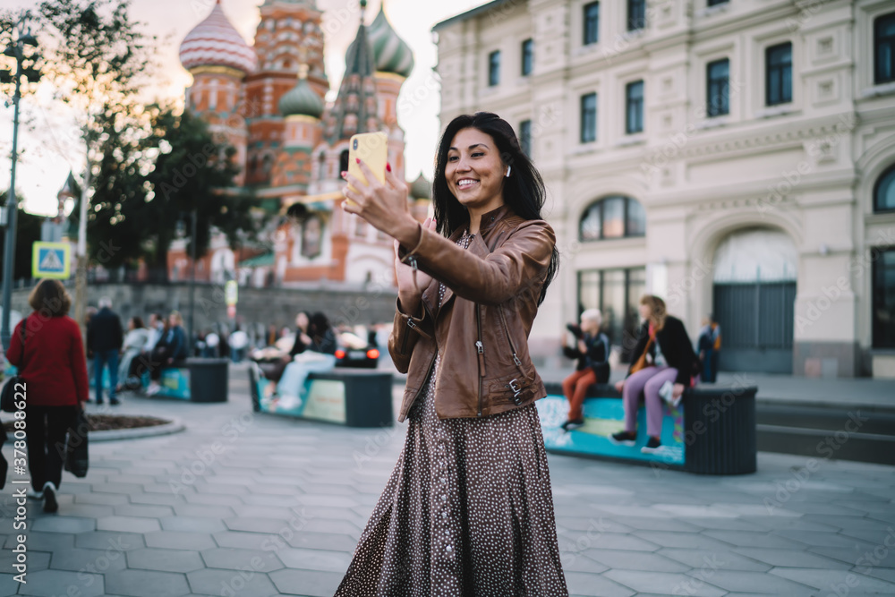 Fototapeta premium Cheerful female travel blogger shooting influence vlog with web sightseeing during solo vacations in Moscow, smiling Asian tourist in earbuds making international video call for communicate in roaming