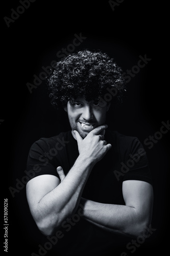 An angry male vampire in black clothes and a wig, bared his teeth, looks at the camera on a black background. Black and white photography. Vertical orientation