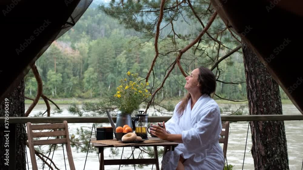 Young Woman in a White Terry Bathrobe Sitting on a Scenic Terrace and Enjoying Breakfast in a Glamping Trip. View from the Inside of a Tent. Luxury Camping, Glamping, Cozy Camping Vacation Lifestyle Young Woman in a White Terry Bathrobe Sitting on a Scenic Terrace and Enjoying Breakfast in a Glamping Trip. View from the Inside of a Tent. Luxury Camping, Glamping, Cozy Camping Vacation Lifestyle