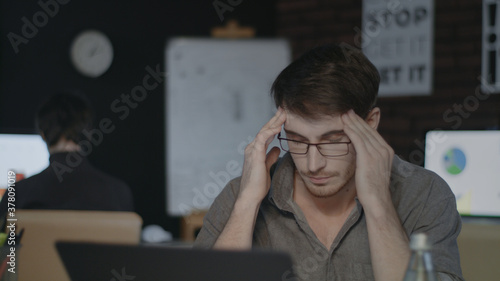 Tired business man touching head in dark office. Sad man working hard on laptop.