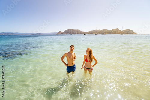 Pareja multiétnica en playa paradisíaca de aguas cristalinas con isla y continente en el fondo, él mirando a cámara y ella a él.