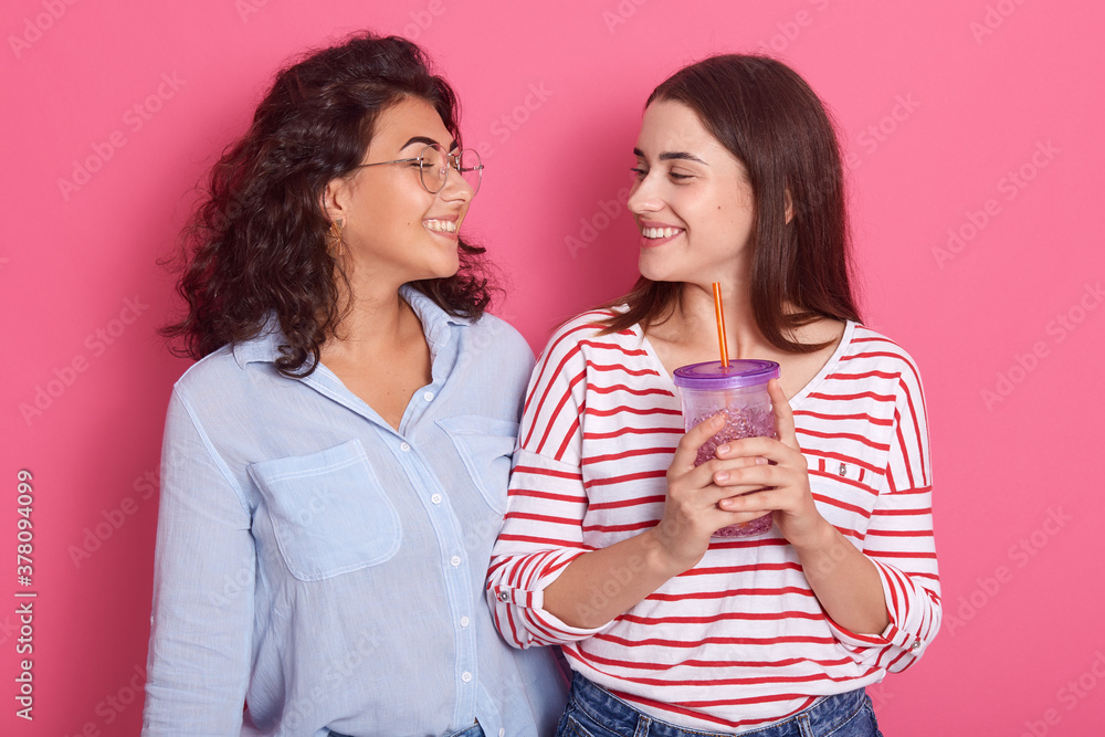 Two girls fun with non alcoholic drink, posing isolated over rosy background, brunette female wearing casual attires, looking smiling at each other. Two girls fun with non alcoholic drink, posing isolated over rosy background, brunette female wearing casual attires, looking smiling at each other.