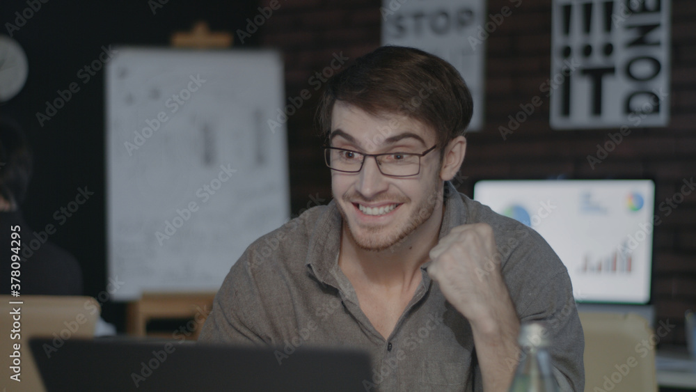 Joyful businessman looking at laptop screen and doing yes gesture in dark office Joyful businessman looking at laptop screen and doing yes gesture in dark office