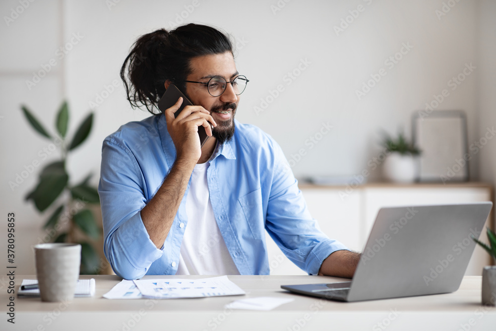 Business Communication. Young Indian Male Entrepreneur Talking On Cellphone While Working On Laptop Business Communication. Young Indian Male Entrepreneur Talking On Cellphone While Working On Laptop