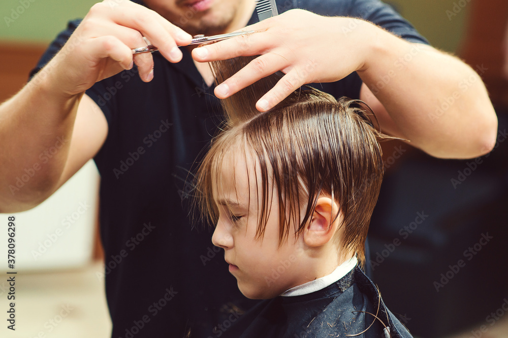 Fototapeta premium Cute little boy getting haircut by hairdresser at the barbershop. Barber man doing kid the hairstyle.