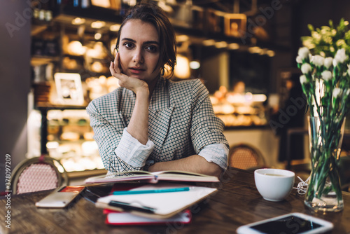 Half length portrait of female entrepreneur creating business plan for small business sitting in own coffee shop and posing, Caucasian student with education notebook looking at camera indoors