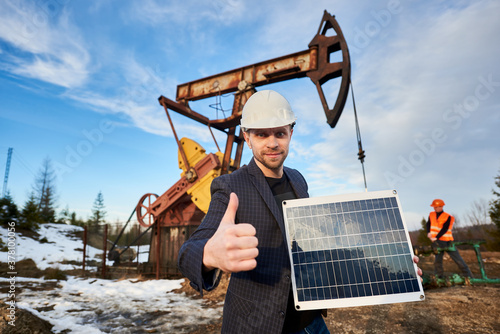 Businessman holding portable solar panel, showing approval gesture. Engineer working with pump jack on territory of oil field on background. Concept of petroleum industry, alternative energy sources.