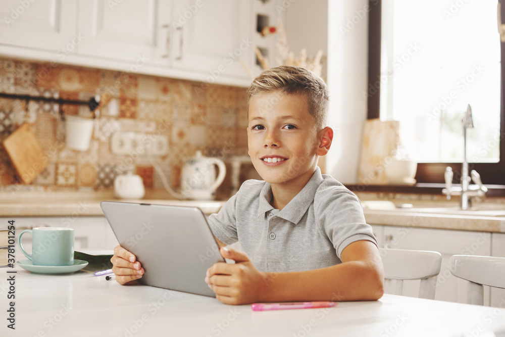 Happy boy sitting with tablet at the table in the home kitchen, chatting on video with friends online Happy boy sitting with tablet at the table in the home kitchen, chatting on video with friends online