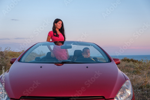 Couple enjoying the sunset from convertible car while the woman is standing and the man sitting